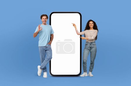 Foto de Un hombre y una mujer se muestran de pie lado a lado de un teléfono con pantalla blanca en blanco, fondo de estudio azul - Imagen libre de derechos