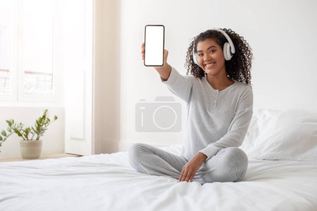 A cheerful Hispanic woman sits cross-legged on a white bed, wearing casual grey clothing and headphones. She is holding up her smartphone facing forward, displaying a blank screen