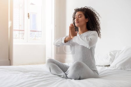 Die hispanische Frau nimmt an einer friedlichen Meditationssitzung teil, auf ihrem Bett sitzend, die Hände in Gebetshaltung zusammengepresst. Das Morgenlicht erfüllt den Raum