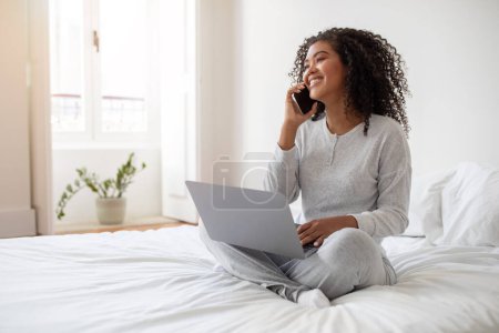 A cheerful young Hispanic woman sits cross-legged on her bed, engaging in conversation on her smartphone. She has a laptop open on her lap, multitasking between a call and her work