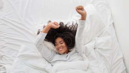 A cheerful young Hispanic woman with curly hair is waking up in a peaceful bedroom. She is stretching her arms and has a content expression on her face, top view, panorama