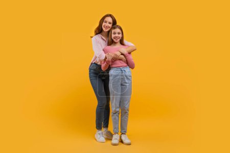 Mutter und Tochter stehen eng beieinander und umarmen einander von hinten. Beide lächeln herzlich und tragen lässig Jeans und helle Oberteile vor leuchtend gelbem Hintergrund..