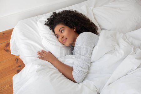 Happy Hispanic woman is laying comfortably in bed, covered by a soft white comforter. Her eyes are open, and she seems to be resting peacefully.