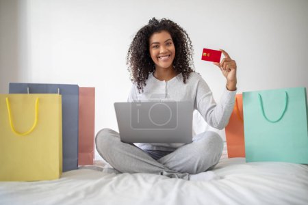 A Hispanic woman shopaholic is seated on a bed, holding a credit card in one hand and a laptop in the other. She appears to be engaged in online shopping or financial transactions.