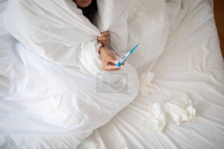 Cropped of woman appears to be sick, tucked under a white blanket, holding a thermometer to check her temperature. Tissues are scattered around her, suggesting she might have a cold or flu
