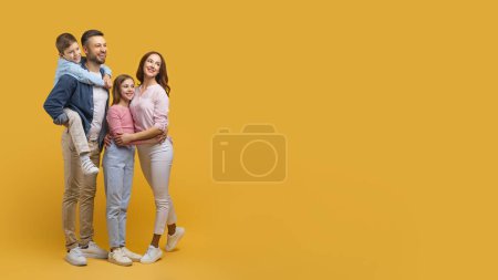 Una familia feliz que consiste en un padre, una madre, un hijo y una hija están muy unidos, abrazándose con sonrisas en sus rostros, mirando el espacio de copia sobre un fondo amarillo