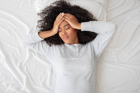 Die hispanische Frau liegt auf dem Rücken liegend im Bett, die Hände auf der Stirn in einer Geste, die auf Kopfschmerzen, Stress, Unwohlsein oder Erschöpfung hindeuten könnte