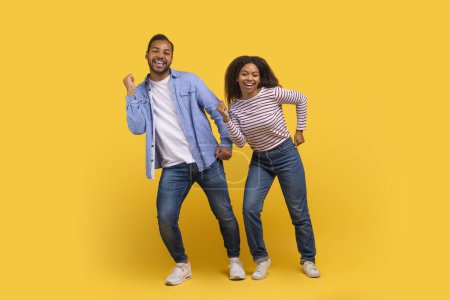 Hombre y mujer afroamericanos están posicionados frente a un sólido telón de fondo amarillo, haciendo movimientos de baile y sonriendo