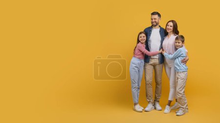 A happy family, consisting of a mother, father, daughter, and son, stand closely together while embracing each other. They are all smiling and looking directly at copy space