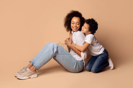 Téléchargez les photos : La jeune femme afro-américaine et son petit fils partagent un moment de tendresse, avec le garçon embrassant amoureusement sa mère par derrière. Ils sont assis sur le sol dans un cadre neutre et neutre - en image libre de droit