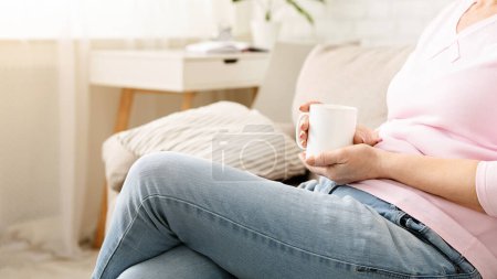 A close-up of woman sitting comfortably on a beige sofa, casually holding a white mug in her hand, with sunlight filtering through the room, cropped