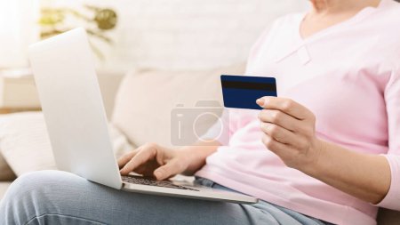 Cropped of woman is seated on a couch, holding a credit card in one hand and a laptop in the other. She appears focused on the laptop screen while balancing the credit card in her hand.