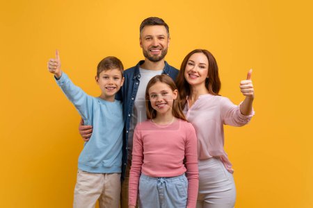 A cheerful family, with two children and their parents, stand close together with bright smiles, each raising their thumb in a sign of approval or success