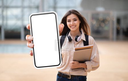 Eine gut gelaunte junge Frau steht draußen und zeigt ein großes Smartphone mit leerem Bildschirm in Richtung Kamera, lässig gekleidet, mit Kopfhörern um den Hals und Notizbüchern im Arm.