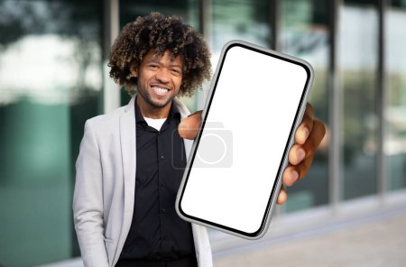 Hombre afroamericano está de pie mientras sostiene un teléfono celular en su mano, mirando a la pantalla. Aparece enfocado y comprometido con el dispositivo, posiblemente enviando mensajes de texto o navegando.