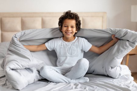 Photo for A cheerful young African American boy sits cross-legged on a bed, spreading a soft blue blanket with both hands in a warmly lit bedroom. - Royalty Free Image