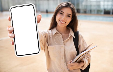 Photo for A woman student is holding a book in one hand and a cell phone in the other. She appears to be reading from the book while glancing at the phone intermittently. - Royalty Free Image