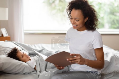 A smiling African American mother is seated on the bed next to her child, who is lying under the covers looking attentive. The mother holds an open book, suggesting that she is reading a story