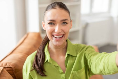 Eine fröhliche junge Frau mit nach hinten gebundenen Haaren macht ein Selfie, ihr Gesicht leuchtet mit einem breiten Lächeln auf. Sie trägt ein grünes Hemd, das ihren strahlenden Teint ergänzt