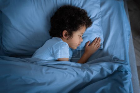 A small African American child is laying comfortably in a bed covered with a blue comforter. Boy appears to be resting peacefully, with eyes closed and hands relaxed by his side