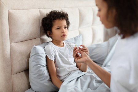 African American young child sits with a contemplative expression on bed while an adult, partially visible, holds out a pill for him to take, likely indicating a moment of healthcare at home.
