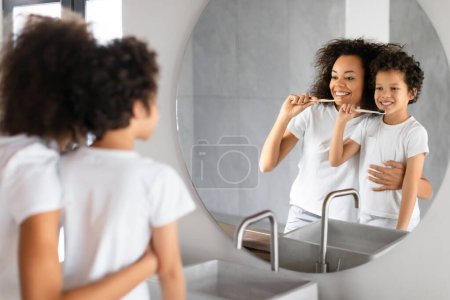 Madre negra y su hijo pequeño se paran frente a un espejo del baño mientras ambos se dedican a su rutina de higiene dental matutina..