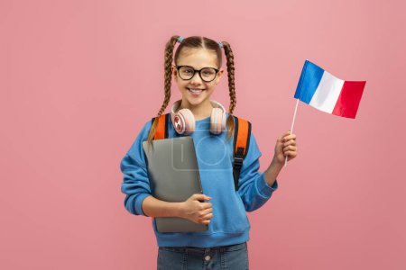 A cheerful young girl with braided hair and glasses is standing against a pink backdrop, holding a small French flag in one hand and a laptop in the other