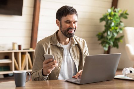 Photo for A bearded man dressed in casual attire sits at a wooden desk in a well-lit, cozy room, engaging with his smartphone in one hand while a laptop is open in front of him - Royalty Free Image