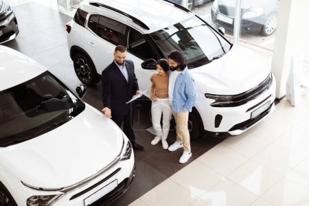 A professional and well-dressed car salesman is engaged in a discussion with Indian couple inside a bright car showroom, high angle view
