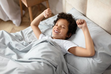 African American young boy is seen lying in bed with his arms extended upward. He looks relaxed and comfortable in his sleep position.
