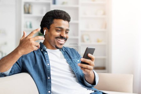 Un homme afro-américain joyeux s'engage dans un appel vidéo, faisant un geste de main comme s'il parlait à quelqu'un tout en se relaxant sur un canapé confortable