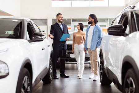 A well-dressed salesman is presenting information to Indian couple in a car dealership showroom. The couple appears interested as they stand between two new vehicles