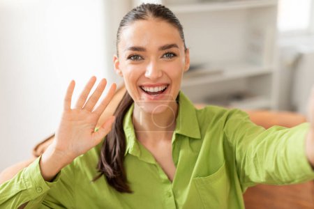 Une jeune femme joyeuse avec les cheveux longs attachés en arrière étend son bras dans une vague, s'engageant dans un appel vidéo décontracté alors qu'elle est vêtue d'un chemisier vert vibrant