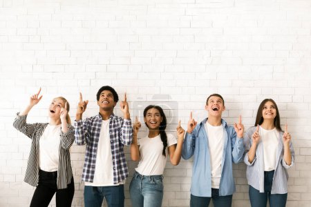 Foto de Un grupo diverso de cinco adolescentes multiétnicos está de pie uno al lado del otro contra una pared de ladrillo blanco, todos sonriendo y mirando hacia arriba mientras apuntan simultáneamente con sus dedos índice - Imagen libre de derechos