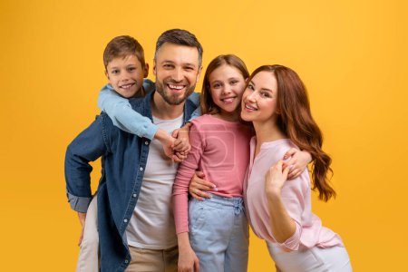 A cheerful family consisting of a father, mother, boy, and girl closely huddled together, sharing a warm embrace while flashing bright smiles against a vibrant yellow backdrop