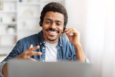 A young African American man with a warm smile is participating in a lively conversation over a video call. He wears a casual denim shirt and effortlessly communicates using his hand gestures