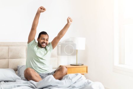 El hombre afroamericano está sentado al borde de una cama, con los brazos levantados en el aire. Parece estar en un estado de celebración o emoción..