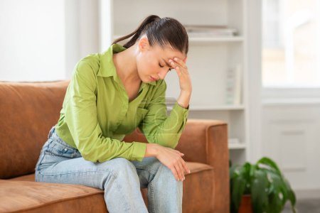 Une femme est assise sur un canapé, la tête dans les mains. Elle semble contemplative ou stressée, profondément dans la pensée ou peut-être éprouver un malaise physique.