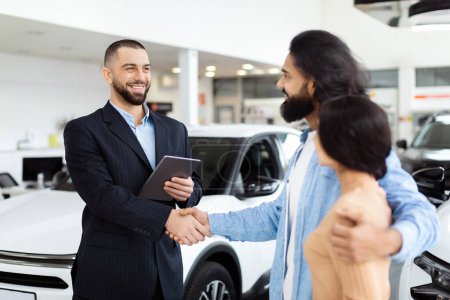 Ein gut gekleideter Verkäufer schüttelt glücklichen indischen Kunden die Hand, vermutlich nachdem er sich auf einen Verkauf geeinigt hat, im Hintergrund ein neues weißes Auto in einem modernen Autohaus-Showroom.