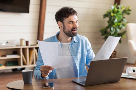 Téléchargez les photos : Un homme barbu avec une expression joyeuse est assis à un bureau en bois, tenant des papiers dans une main avec un ordinateur portable ouvert devant lui - en image libre de droit