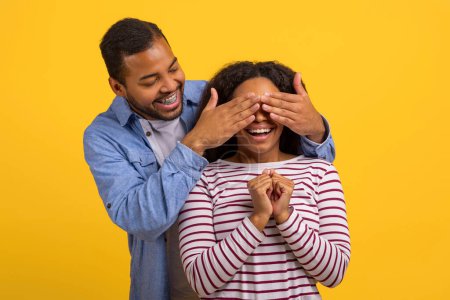 A smiling young African American man is standing behind a woman, playfully covering her eyes with his hands as she reacts with a joyful expression of surprise and anticipation