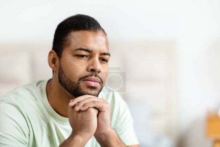 El hombre afroamericano aparece profundamente en el pensamiento mientras apoya su barbilla en su mano, su mirada ligeramente fuera de cámara. Su expresión facial transmite contemplación o preocupación, espacio de copia