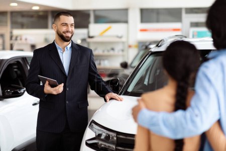 A professional salesman, clad in a smart suit, stands by a shiny new car with a digital tablet in hand, confidently explaining features to an interested Indian couple in a car dealership showroom