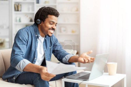 Ein fröhlicher afroamerikanischer Mann sitzt gemütlich auf einem Sofa und nutzt einen Laptop für ein virtuelles Treffen. Er trägt Kopfhörer und hält ein Dokument in der Hand