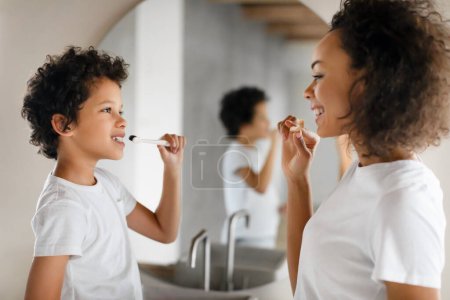 African American woman is brushing her teeth next to a little boy. They are standing in front of a bathroom sink. The woman holds a toothbrush while the little boy watches intently.