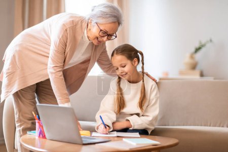 Grandmother standing by little girl granddaughter drawing and looking at laptop screen, doing homework, home interior, copy space
