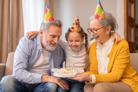 A young girl wearing a party hat shares a cheerful moment with her beaming grandparents as they sit on a couch together, with the girl holding a small birthday cake adorned with lit candles.