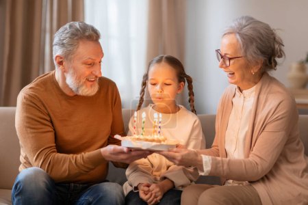 A young girl sitting between her smiling grandparents is blowing out candles on a birthday cake that her grandfather is holding. They appear to be seated comfortably in a living room