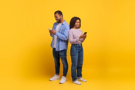 Hombre y mujer afroamericanos están de pie con sus espaldas tocando, cada uno absorbido en su propio teléfono inteligente. Están casualmente vestidos y parecen estar disfrutando de un momento de conectividad.