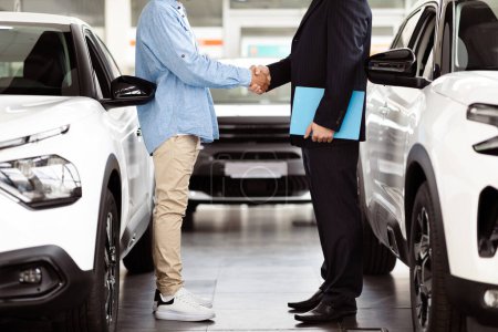 Téléchargez les photos : Cropped of two men are standing inside a bright and spacious car showroom. They are shaking hands with each other, appearing satisfied and confident after finalizing a deal on a new vehicle. - en image libre de droit